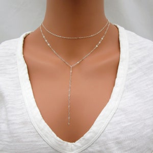 May include: A silver layered necklace with a long, delicate chain extending from the shorter chain. The necklace is worn on a model with a white shirt.