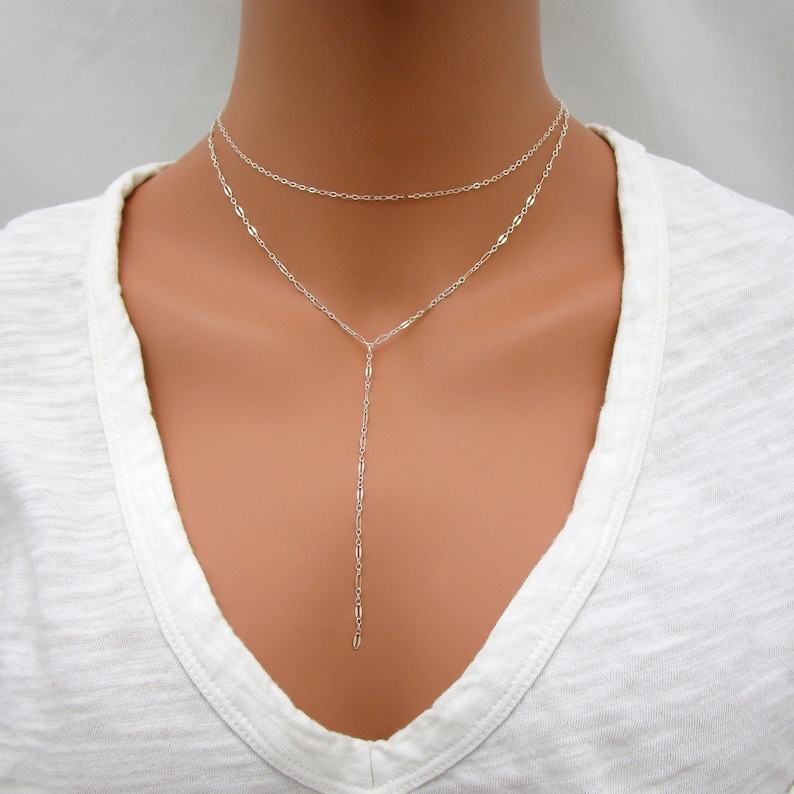May include: A silver layered necklace with a long, delicate chain extending from the shorter chain. The necklace is worn on a model with a white shirt.