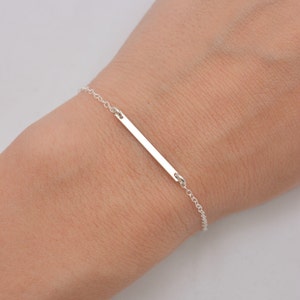 May include: A silver bar bracelet with a delicate chain. The bar is simple and sleek, making it a minimalist and elegant piece of jewelry.