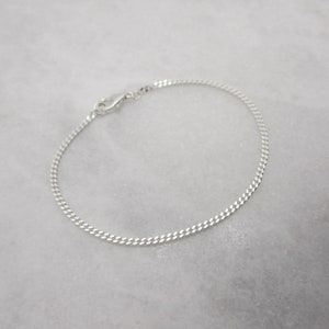K&ouml;nnte beinhalten: Ein zartes Armband aus Silberkette mit einem Karabinerverschluss. Das Armband hat ein flaches Panzerketten-Design, das Licht reflektiert. Der Schmuck wird auf einer wei&szlig;en Marmoroberfl&auml;che pr&auml;sentiert.