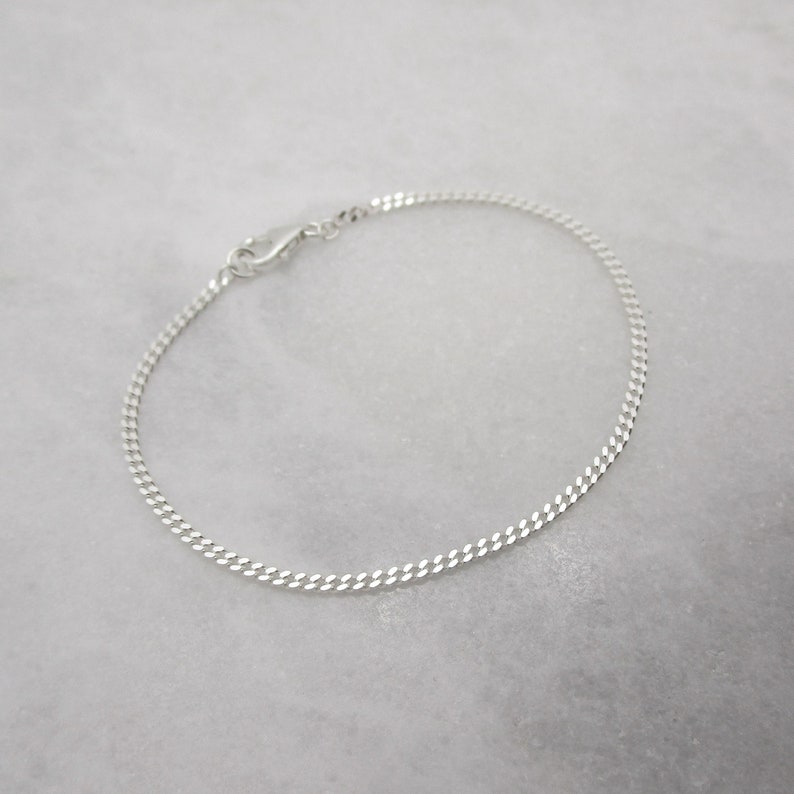 K&ouml;nnte beinhalten: Ein zartes Armband aus Silberkette mit einem Karabinerverschluss. Das Armband hat ein flaches Panzerketten-Design, das Licht reflektiert. Der Schmuck wird auf einer wei&szlig;en Marmoroberfl&auml;che pr&auml;sentiert.
