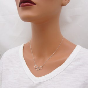 Pode incluir: Um colar de prata com um pingente de tr&ecirc;s c&iacute;rculos entrela&ccedil;ados. O colar est&aacute; em um manequim branco que est&aacute; vestindo uma camiseta branca.