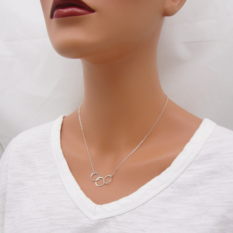 Pode incluir: Um colar de prata com um pingente de tr&ecirc;s c&iacute;rculos entrela&ccedil;ados. O colar est&aacute; em um manequim branco que est&aacute; vestindo uma camiseta branca.