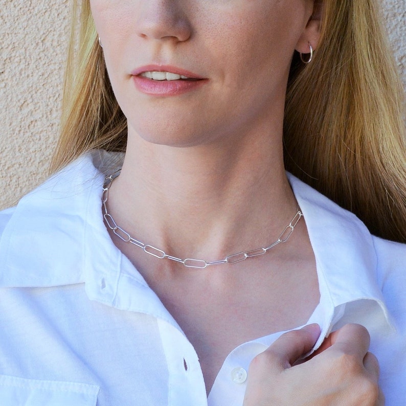 May include: A silver chain necklace with elongated oval links. The necklace is worn over a white button-down shirt. The model is wearing a pair of small silver hoop earrings. The background is a neutral beige color.