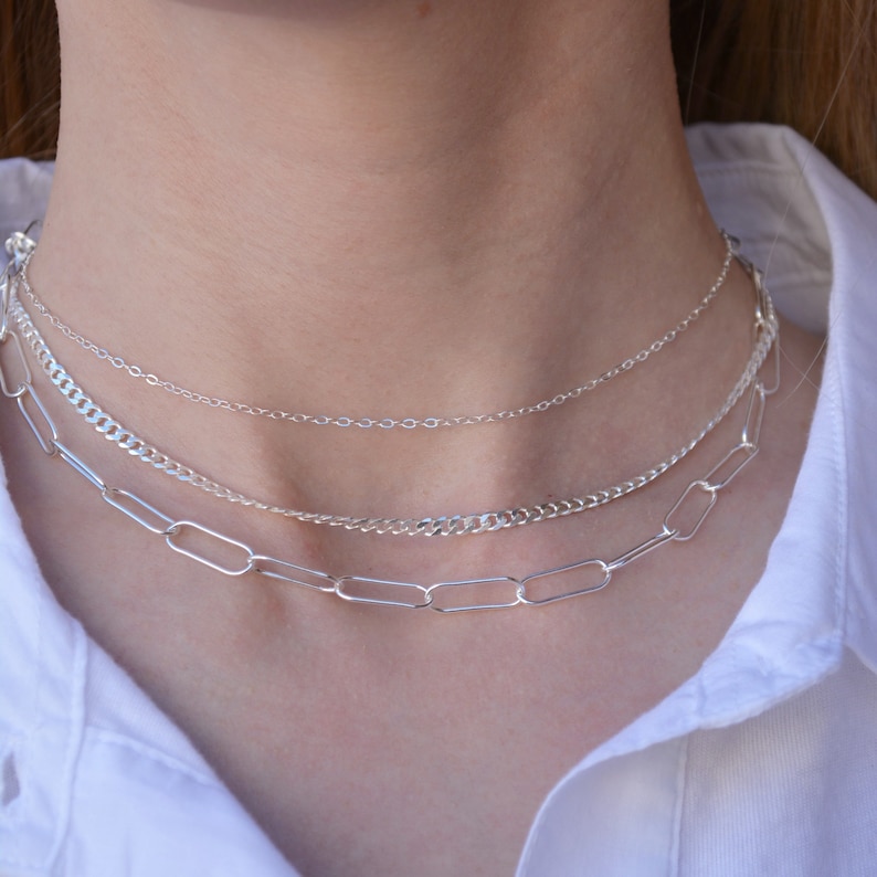 May include: A layered silver necklace set. The necklaces include a delicate chain, a curb chain, and a chain with elongated oval links. The jewelry is displayed against a white shirt.