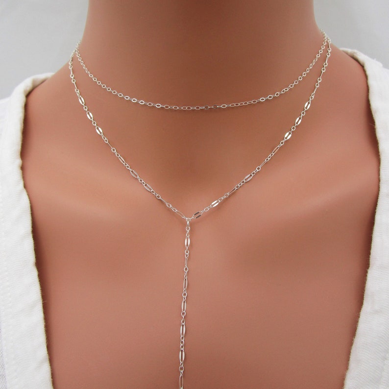 May include: A silver layered necklace with a delicate chain and a longer, dangling chain. The necklace is worn on a model's neck.
