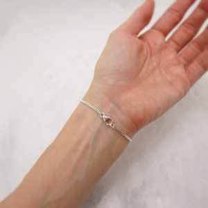 K&ouml;nnte beinhalten: Ein filigranes Silberarmband mit einem Curb-Kettendesign. Das Armband wird mit einem Karabinerverschluss gesichert. Der Schmuck wird an einem Handgelenk gezeigt.