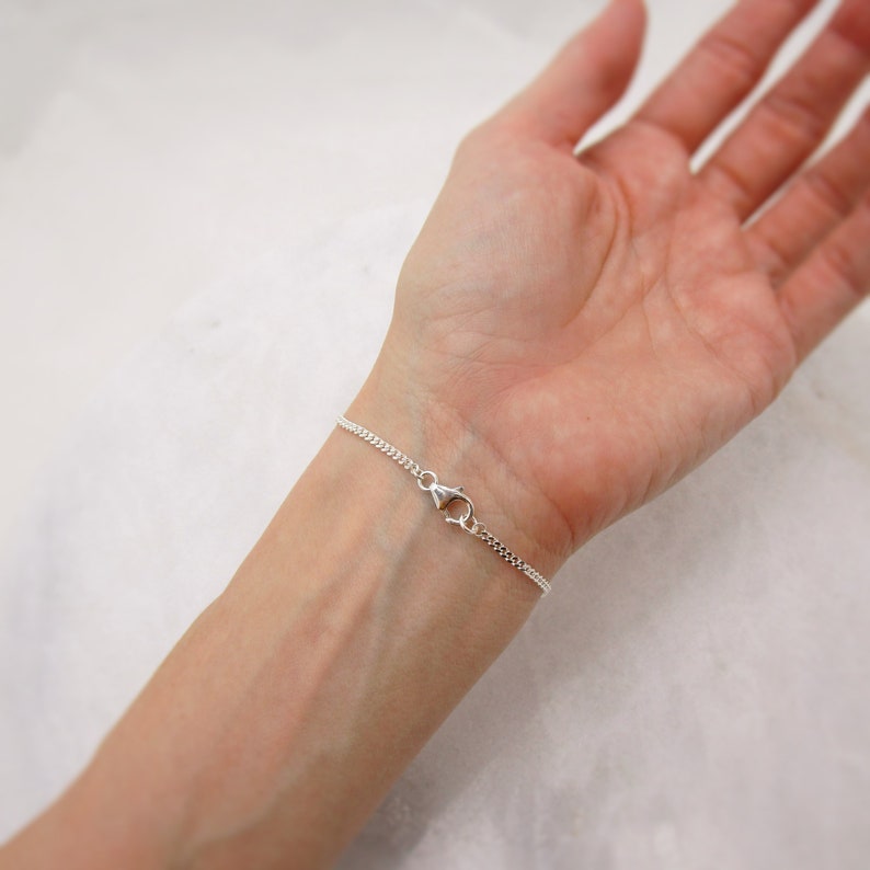 K&ouml;nnte beinhalten: Ein filigranes Silberarmband mit einem Curb-Kettendesign. Das Armband wird mit einem Karabinerverschluss gesichert. Der Schmuck wird an einem Handgelenk gezeigt.