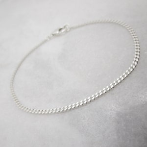 K&ouml;nnte beinhalten: Ein zartes Armband aus Silberkette mit einem Karabinerverschluss. Das Armband hat ein flaches Panzerketten-Design. Der Schmuck wird auf einer wei&szlig;en Marmoroberfl&auml;che pr&auml;sentiert.