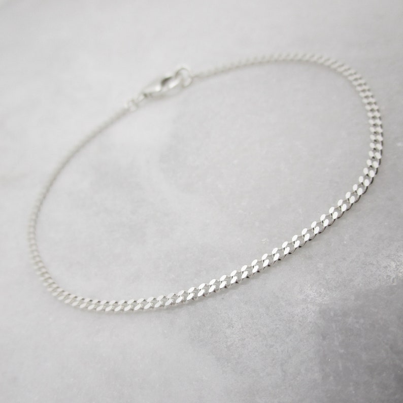K&ouml;nnte beinhalten: Ein zartes Armband aus Silberkette mit einem Karabinerverschluss. Das Armband hat ein flaches Panzerketten-Design. Der Schmuck wird auf einer wei&szlig;en Marmoroberfl&auml;che pr&auml;sentiert.