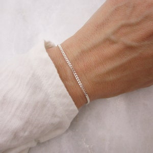 K&ouml;nnte beinhalten: Ein filigranes Silberarmband. Das Armband hat kleine, ineinandergreifende Glieder. Das Armband wird am Handgelenk getragen, vor einem wei&szlig;en Langarmhemd. Die Kette ist etwa 18 cm lang.