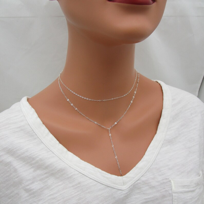 May include: A silver layered necklace with a delicate chain and a longer, dangling chain. The necklace is worn on a model with a white shirt.