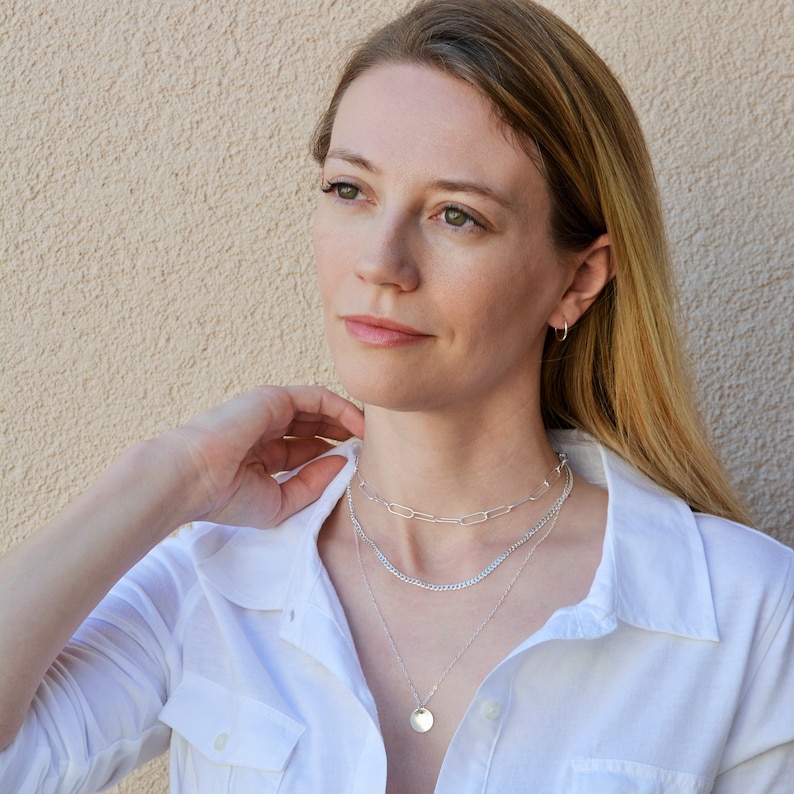 May include: A woman wearing a white shirt and three silver necklaces. The necklaces include a chain link, a curb chain, and a pendant necklace with a small, round disc. She is also wearing small silver hoop earrings.