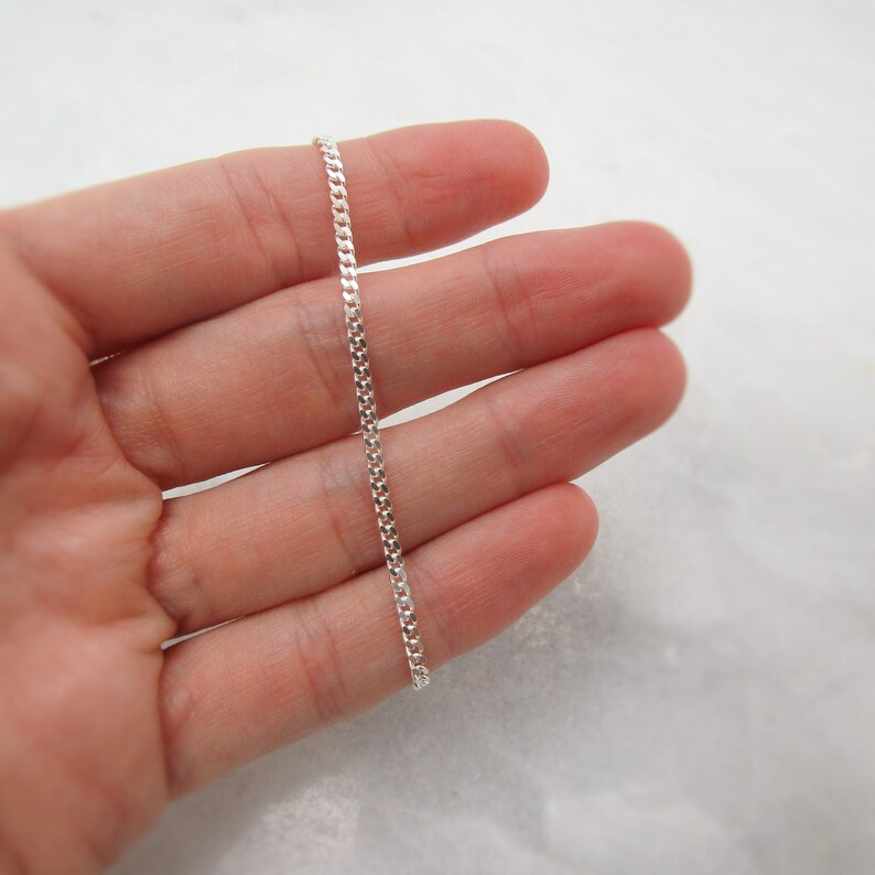 K&ouml;nnte beinhalten: Eine zarte Silber-Kettenkette, gehalten in einer Hand. Die Kette besteht aus kleinen, ineinandergreifenden Gliedern, die eine subtile, reflektierende Oberfl&auml;che erzeugen. Die Kette wird vor einem weichen, neutralen Hintergrund gezeigt.