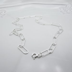 May include: A delicate silver chain necklace featuring a series of elongated oval links. The necklace has a simple, modern design with a secure clasp closure. The chain is displayed on a light gray surface.