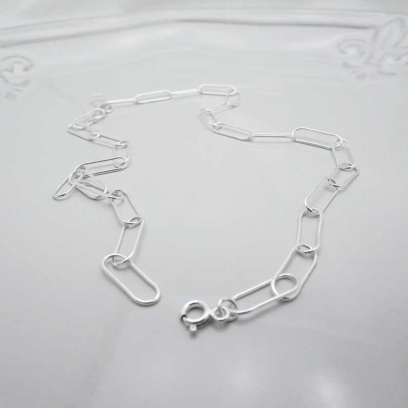 May include: A delicate silver chain necklace featuring a series of elongated oval links. The necklace has a simple, modern design with a secure clasp closure. The chain is displayed on a light gray surface.