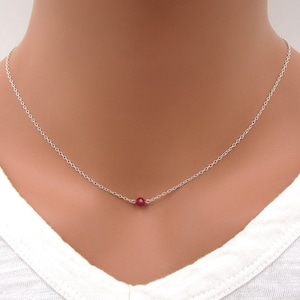May include: A delicate silver chain necklace with a single, small, red gemstone bead.