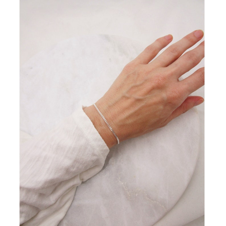 K&ouml;nnte beinhalten: Ein filigranes Silberarmband. Das Armband hat ein schlichtes, elegantes Design mit kleinen, miteinander verbundenen Gliedern. Es wird an einem Handgelenk vor wei&szlig;em Hintergrund getragen. Ein klassisches Schmuckst&uuml;ck.