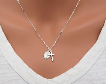 Pode incluir: Um colar de tom prateado com uma corrente fina e dois pequenos pingentes. Um pingente é uma cruz simples e lisa, e o outro é uma medalha oval com uma figura religiosa em relevo, que repousa sobre uma peça de vestuário branca.