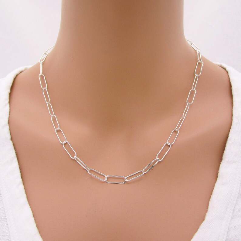 May include: A silver-colored chain necklace with elongated oval links. The necklace is worn around the neck, resting on a white shirt. The links are evenly spaced, creating a simple, elegant design.