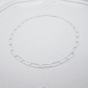 May include: A delicate silver chain necklace featuring a series of elongated oval links. The necklace is displayed on a white surface, showcasing its simple yet elegant design. The chain is approximately 16 inches in length.