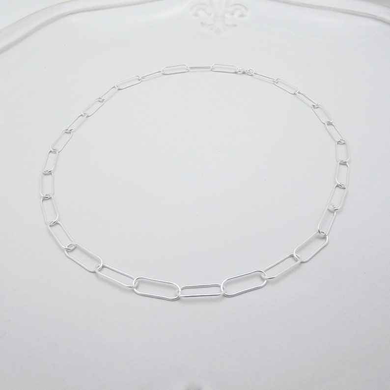 May include: A delicate silver chain necklace featuring a series of elongated oval links. The necklace is displayed on a white surface, showcasing its simple yet elegant design. The chain is approximately 16 inches in length.