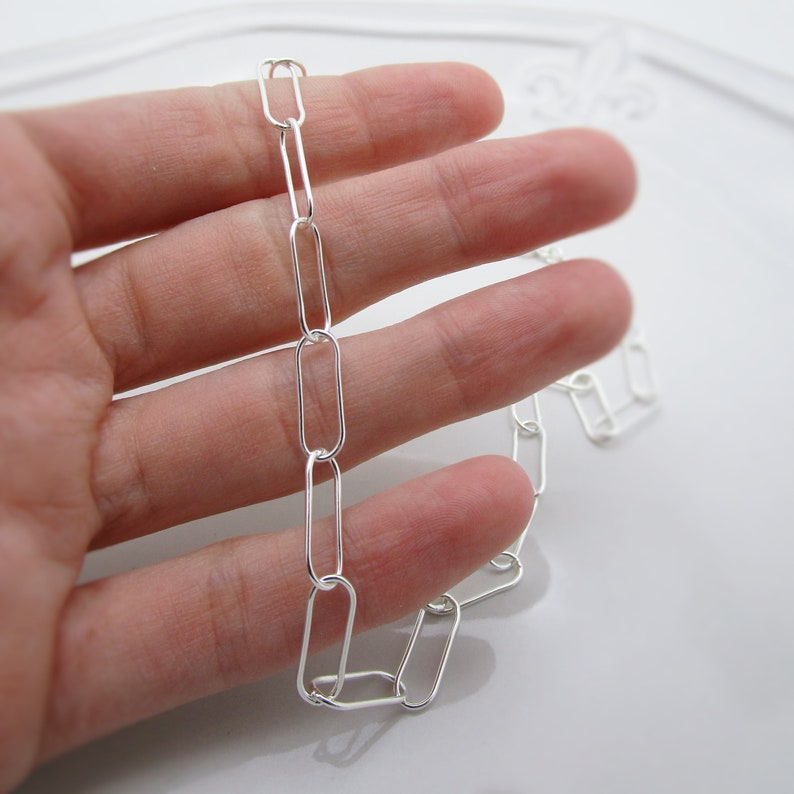 May include: A silver chain necklace with elongated oval links. The chain is held in a hand, showcasing the bright, reflective metal. The links are evenly spaced, creating a simple yet elegant design. The necklace is a delicate piece of jewelry.