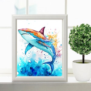 Puede incluir: Una pintura de acuarela de un tibur&oacute;n saltando fuera del agua. El tibur&oacute;n es azul, naranja y blanco. El agua es azul y verde. Hay salpicaduras de pintura rosa y amarilla alrededor del tibur&oacute;n.