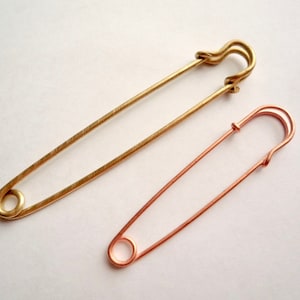 May include: Two safety pins, one gold and one copper, are shown against a white background. The gold safety pin is larger than the copper safety pin.
