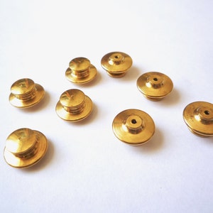 May include: Eight gold-colored metal pin backs. The pin backs are round with a raised center and a flat bottom.