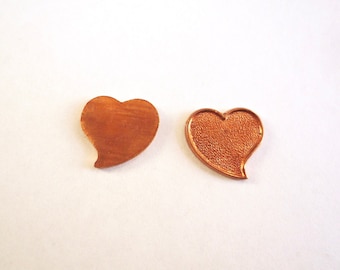 May include: Two heart-shaped copper-colored metal blanks. One is a solid heart shape, and the other has a textured surface with a raised edge, designed to hold an insert. Both are on a white background.