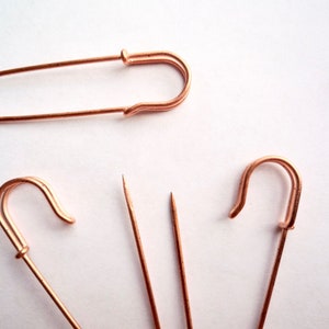 May include: Four rose gold safety pins, two are standard size and two are smaller. The larger pins are open and the smaller pins are closed.
