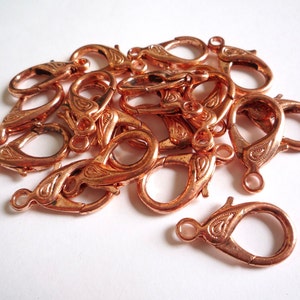 May include: A pile of copper-colored lobster clasps with a decorative design. The clasps are used for jewelry making.