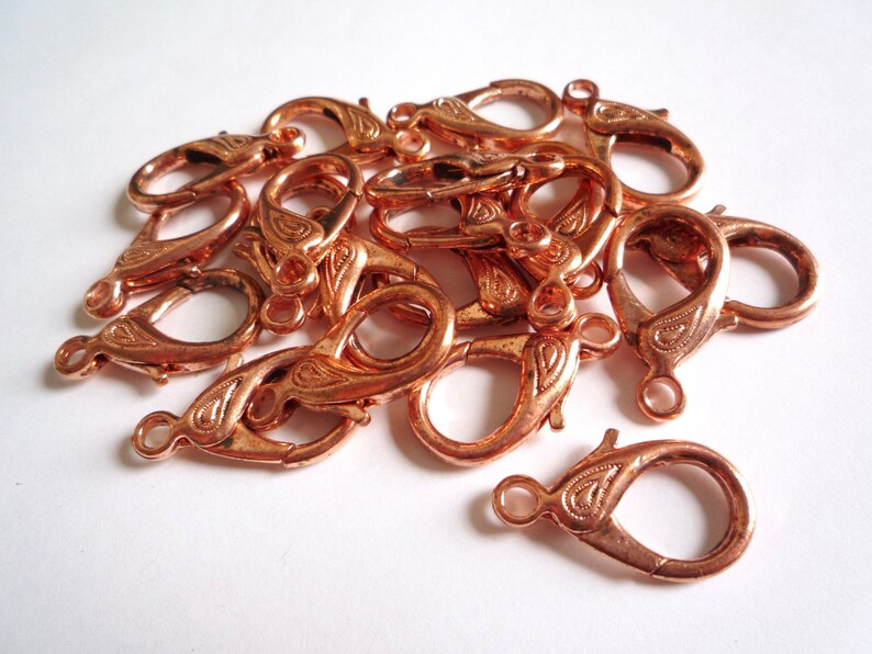 May include: A pile of copper-colored lobster clasps with a decorative design. The clasps are used for jewelry making.
