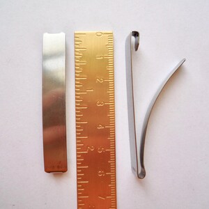 Op de afbeelding: Drie metalen stukken, twee zijn plat en &eacute;&eacute;n is gebogen. De platte stukken zijn zilverkleurig en het gebogen stuk is zilverkleurig met een gouden liniaal erachter.