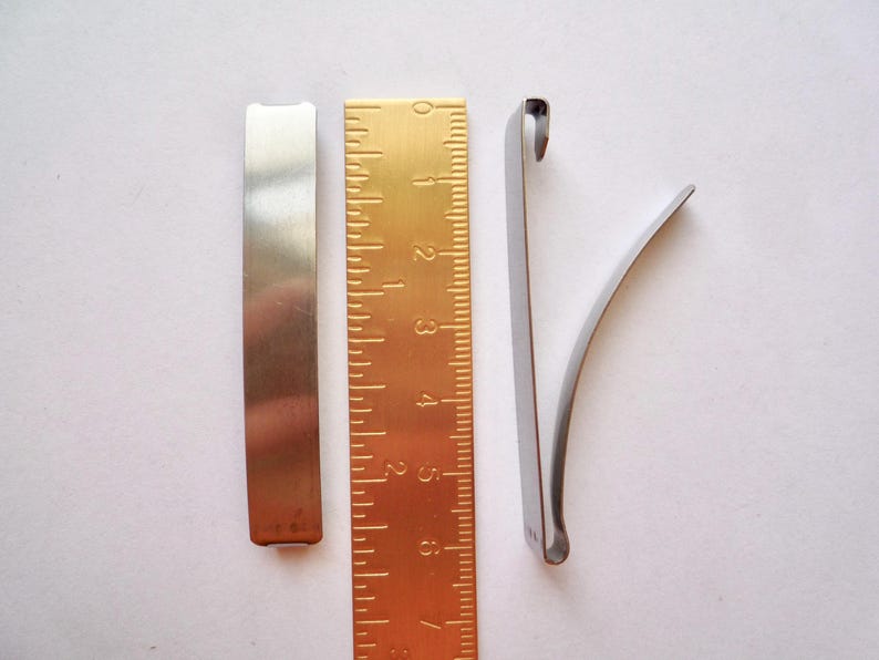 Op de afbeelding: Drie metalen stukken, twee zijn plat en &eacute;&eacute;n is gebogen. De platte stukken zijn zilverkleurig en het gebogen stuk is zilverkleurig met een gouden liniaal erachter.