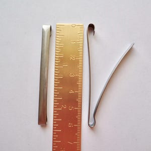 Op de afbeelding: Drie zilveren metalen haarclips, &eacute;&eacute;n recht en twee gebogen, naast een gouden liniaal met markeringen van 1 tot 6.