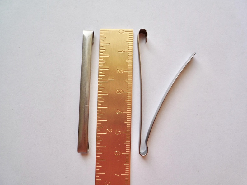 Op de afbeelding: Drie zilveren metalen haarclips, &eacute;&eacute;n recht en twee gebogen, naast een gouden liniaal met markeringen van 1 tot 6.