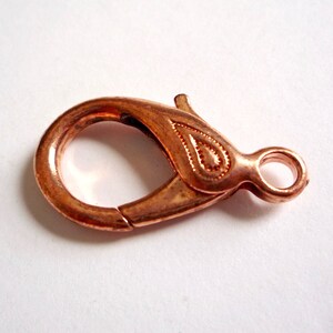 May include: A copper-colored lobster clasp with a decorative design. The clasp is a metal fastener used for jewelry making.
