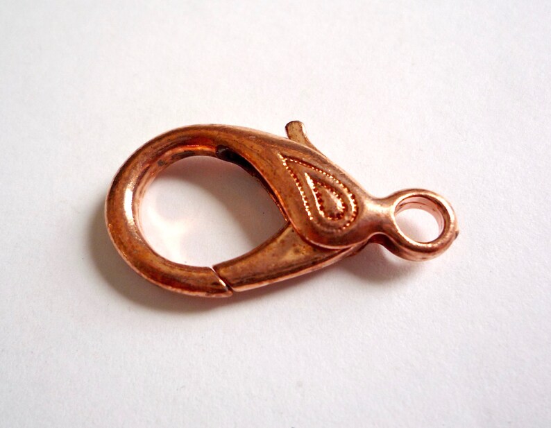 May include: A copper-colored lobster clasp with a decorative design. The clasp is a metal fastener used for jewelry making.