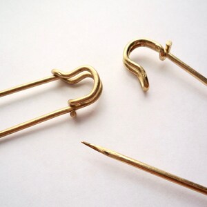 May include: Two gold safety pins, one open and one closed, on a white background.