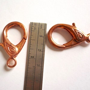 May include: Two copper-colored lobster clasps with a decorative design. The clasps are approximately 1 inch in length.