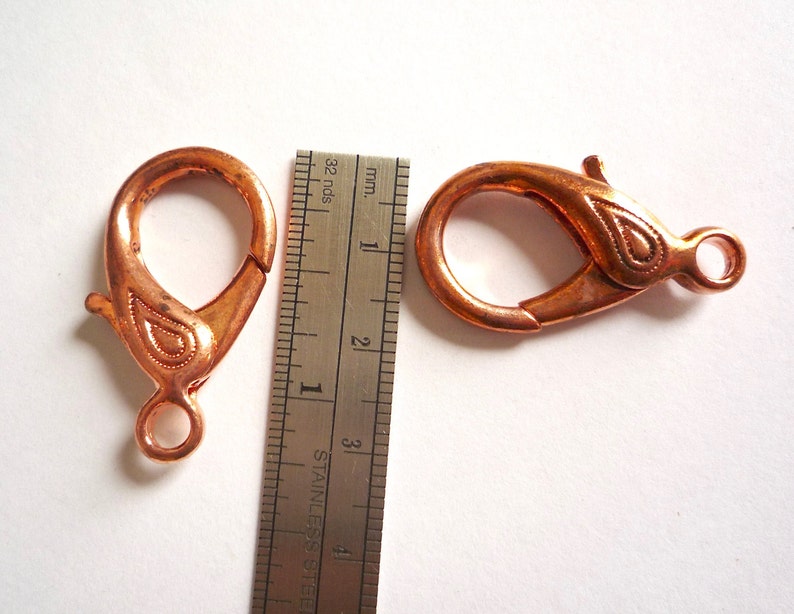 May include: Two copper-colored lobster clasps with a decorative design. The clasps are approximately 1 inch in length.
