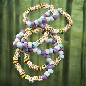 Op de afbeelding: Meerdere kleurrijke armbanden met de woorden "WANNA GET HIGH" gespeld in witte letterkralen. De armbanden hebben een mix van paarse, lichtblauwe en witte kralen, met accenten van geel, rood en groen.