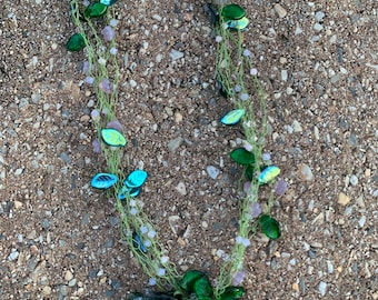 May include: A handmade necklace featuring a delicate design with green netting, iridescent leaf-shaped beads, and small purple beads. The necklace includes a cluster of purple flowers and green leaves at the bottom. A small tag reads "Handmade with love."