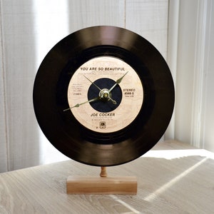 May include: A black vinyl record clock with a gold clock face. The record label features the song "You Are So Beautiful" by Joe Cocker. The clock is on a wooden stand.
