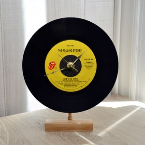 May include: A black vinyl record clock featuring a yellow label with "THE ROLLING STONES" and "SHE'S SO COLD" printed on it. The clock has gold-colored hands and is mounted on a small wooden stand. The Rolling Stones logo is visible.