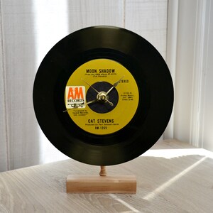 May include: A circular wall clock crafted from a black vinyl record featuring a yellow A&M Records label for Cat Stevens' Moon Shadow. The clock has gold-colored metal hands and is mounted on a rectangular light-colored wooden base stand.