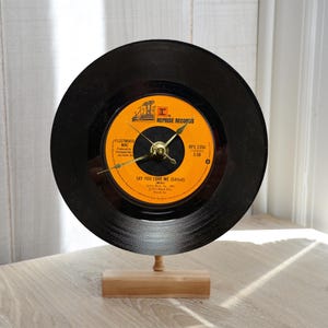 May include: A unique clock made from a black vinyl record. The clock face features an orange label with the text "FLEETWOOD MAC" and "SAY YOU LOVE ME (Edited)". The clock has gold-colored hands and is mounted on a wooden stand.