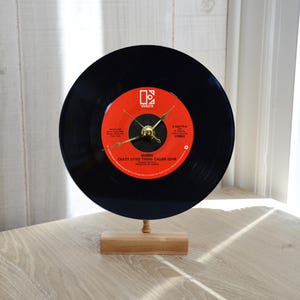 May include: A black vinyl record clock featuring the Queen song "Crazy Little Thing Called Love". The clock is mounted on a wooden stand and has gold hands.